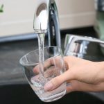 Hand holding a glass under a kitchen faucet, filling it with clean drinking water, highlighting the importance of backflow prevention in maintaining water quality and safety.